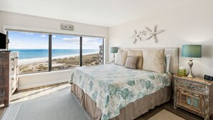 Et soveværelse med et stort vindue der giver udsigt til stranden, en seng med et sengetæppe med et tropisk blad-mønster, en søstjerne som dekoration på væggen, og et rustikt natbord med en lampe.