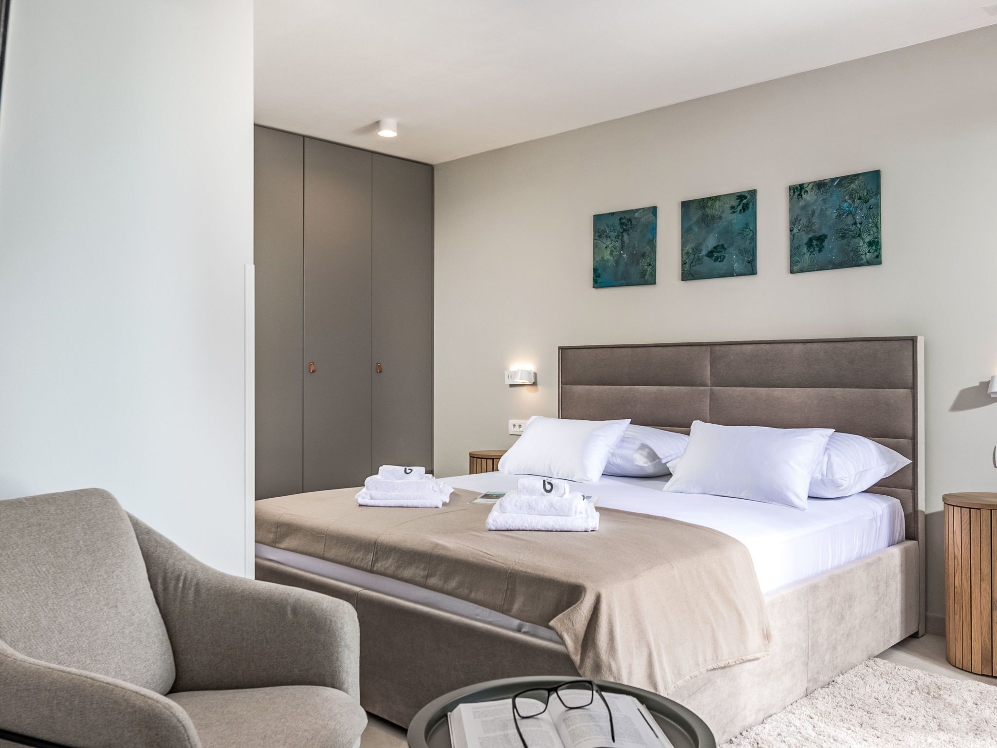 A modern hotel room with a large bed, a sofa, and a small table with glasses on it. There are three paintings on the wall and a wardrobe in the corner.
