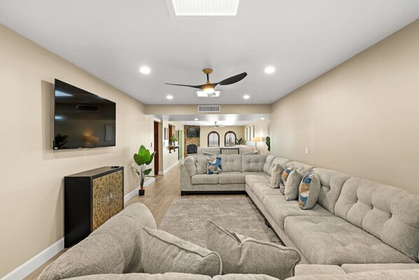 A spacious living room with a large sectional sofa, a flat-screen TV mounted on the wall, and a ceiling fan.