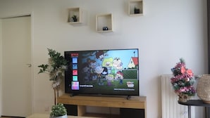A television displaying a cartoon, with a Christmas tree and a small plant on a wooden stand.