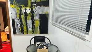 Ein kleiner Raum mit einem Gemälde an der Wand, einem runden Tisch mit zwei Stühlen, einem Fenster mit Jalousien und einem roten Teppich.