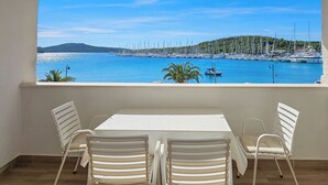 Un balcón con mesa y sillas blancas, con vista a un puerto deportivo con barcos y una colina distante.