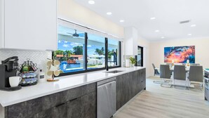 A modern kitchen with a large window, a coffee machine, and a view of a pool and buildings. - Waterfront | Boat Rental | Heated Pool | Games Room | Sleeps 8 (Lighthouse Point)