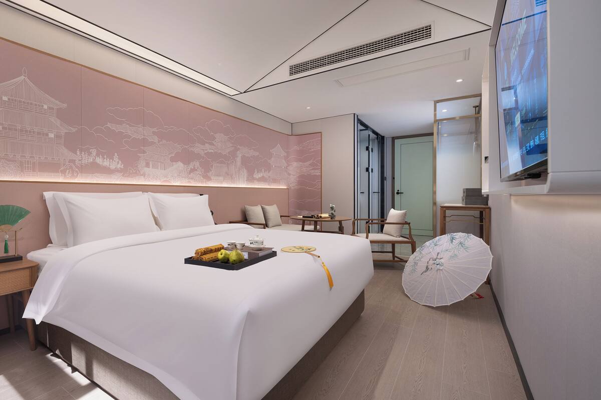 A hotel room with a large bed, bedside table with a lamp, a small table with a teapot, a chair, and a TV mounted on the wall.