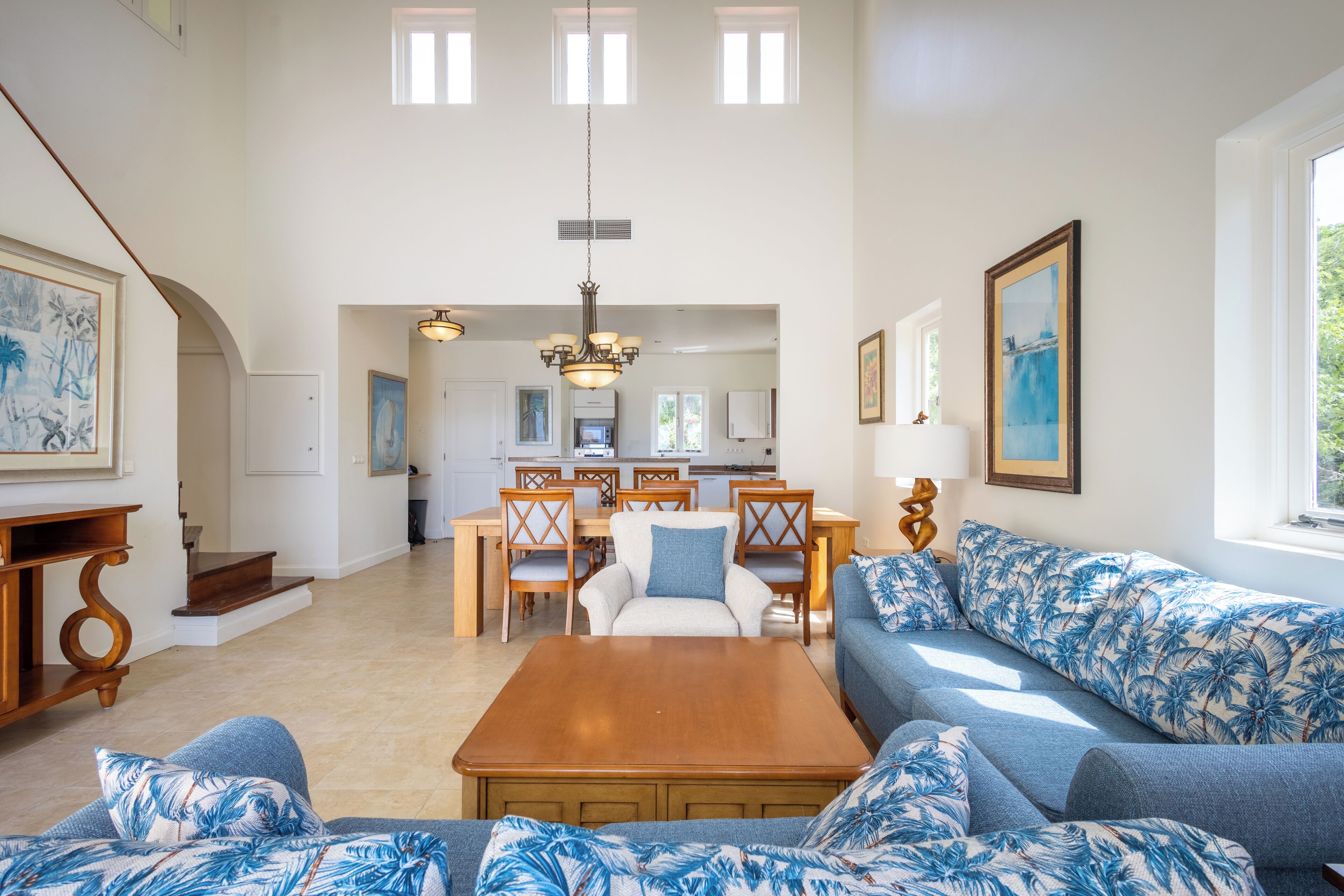 Un salon spacieux avec une table basse en bois, un canapé bleu à motifs tropicaux et un coin repas comprenant des chaises en bois et un lustre.
