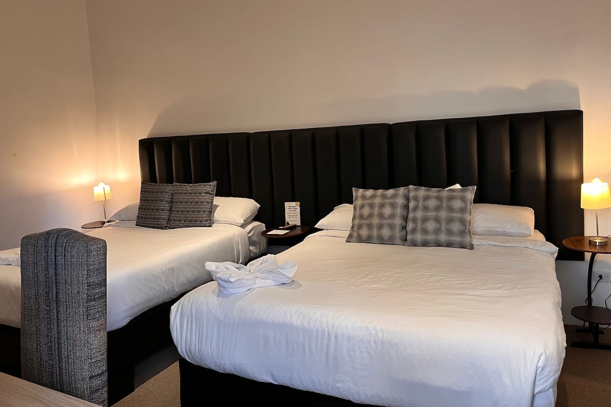 Hotellihuone, jossa on kaksi sänkyä. Jokaisessa sängyssä on valkoiset lakanat ja tyynyt, tumma pääty sekä yöpöydät lampuineen ja pieni pöytä lampun kanssa.