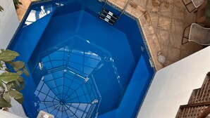 Una bañera hexagonal con techo de cúpula de vidrio, rodeada de muros de piedra y una escalera de metal.