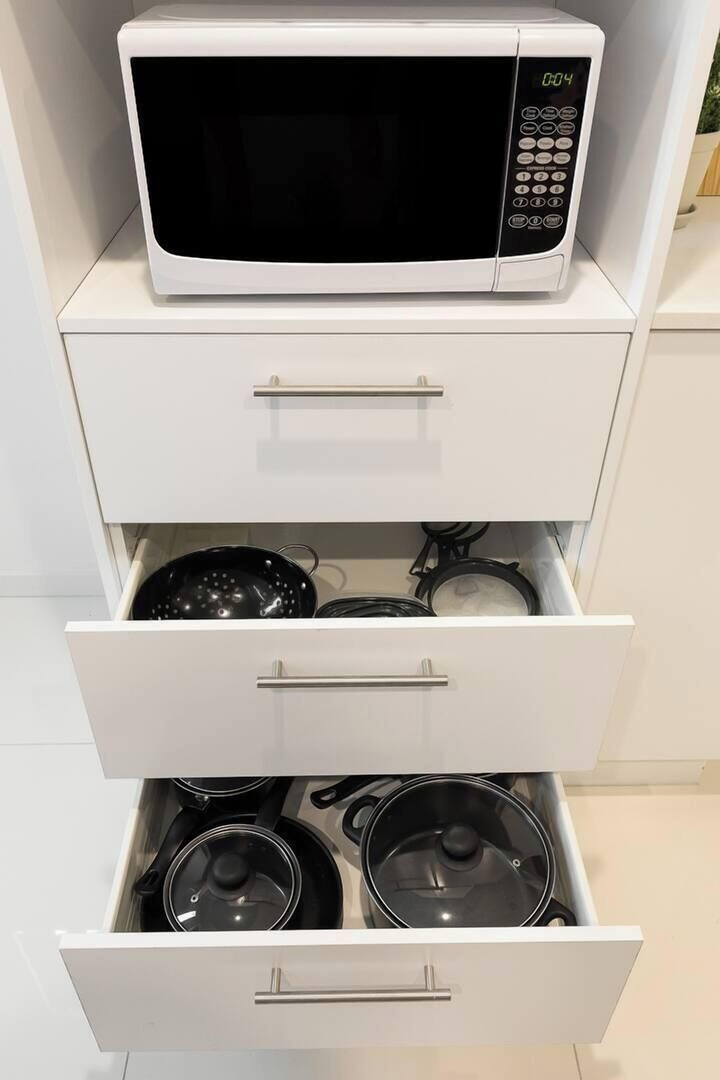 A kitchen with a microwave on top of a white cabinet, a drawer below it containing pots, and a lower drawer with more pots.