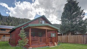 A wooden cabin with a porch, surrounded by a fenced yard and a forested backdrop.