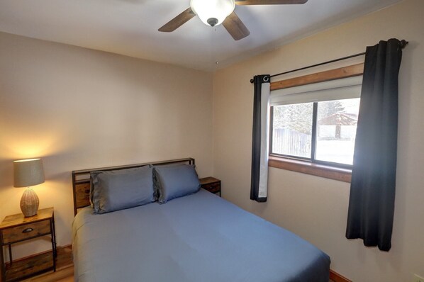 A bedroom with a bed, a nightstand, a lamp, a ceiling fan, a window with curtains, and a view of the outside. - River's Edge Retreat (Red River)