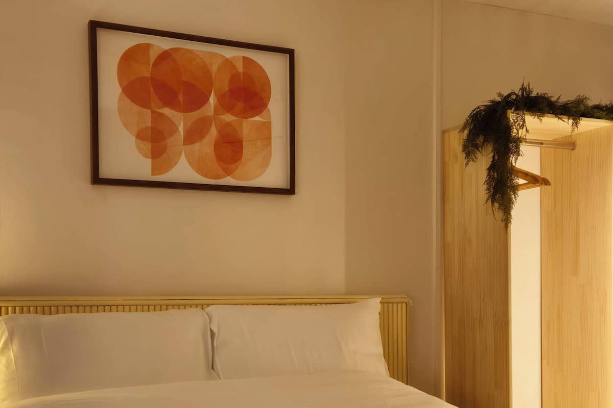 A neatly made bed with white linens, a framed abstract painting above it, and a wooden door with a green plant hanging on it.