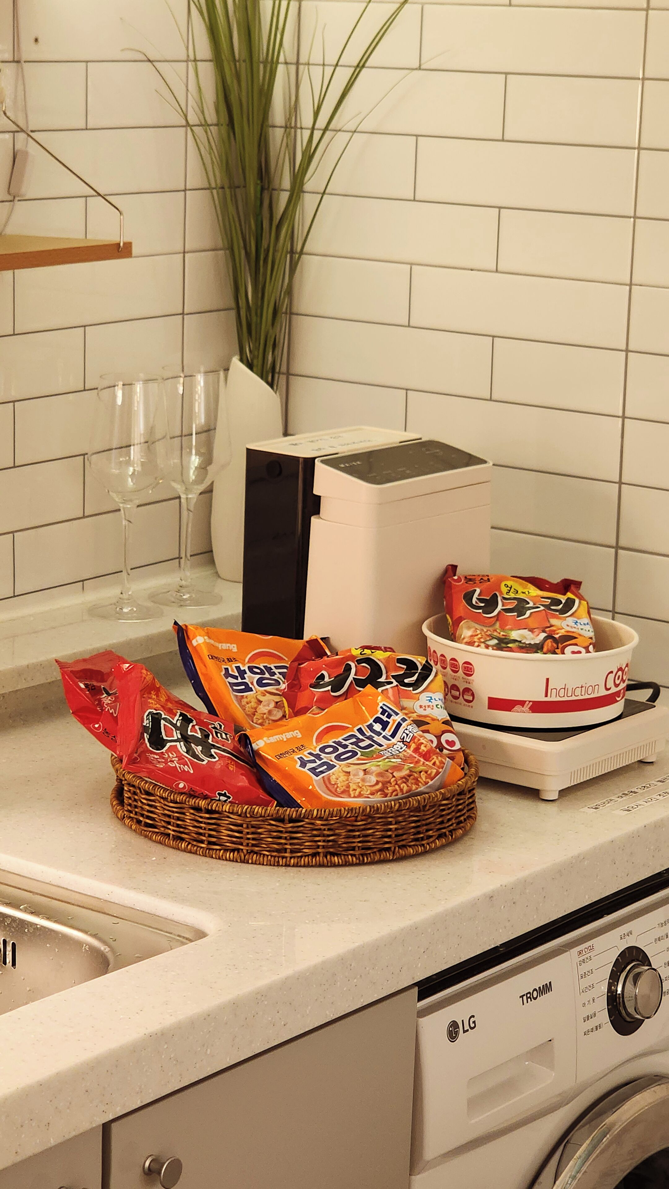 A kitchen counter with a washing machine, a basket of instant noodles, and a few wine glasses.
