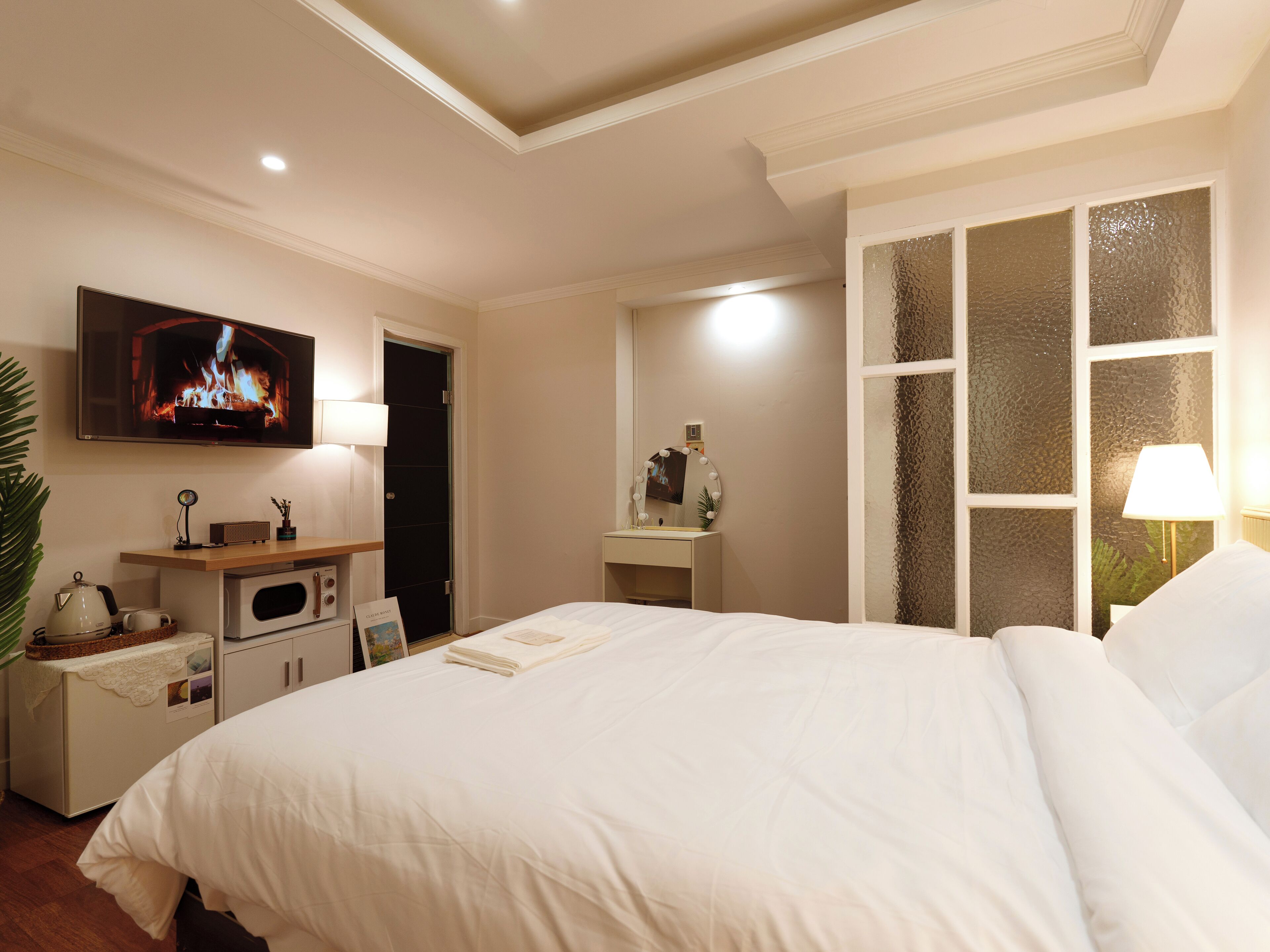 Une chambre d’hôtel dotée d’un grand lit, d’un téléviseur fixé au mur, d’un bureau avec une lampe, d’une coiffeuse avec un miroir et d’une fenêtre ornée de panneaux de verre décoratifs.