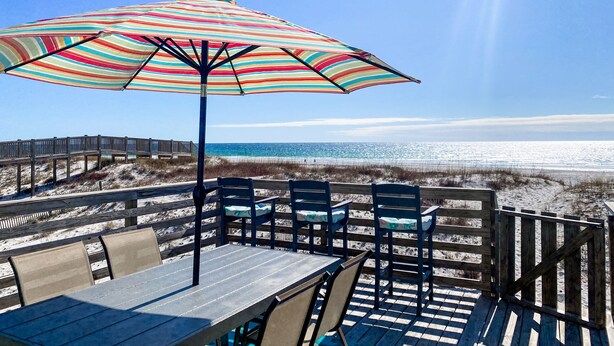Una terraza de madera con una mesa, sillas y una sombrilla, con vista a la playa y al océano.