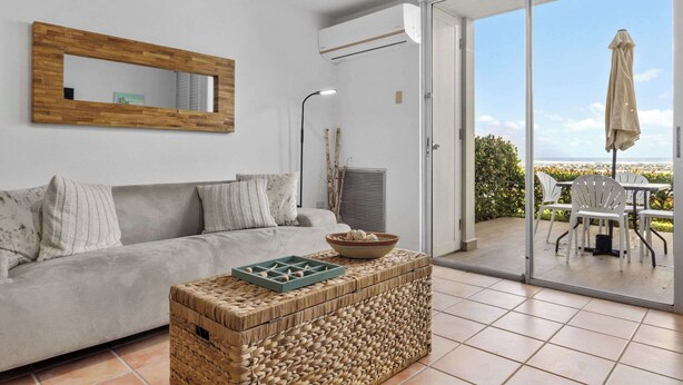 Ein Wohnzimmer mit einer Couch, einem gewebten Couchtisch, einem Spiegel und einer Schiebetür zu einer Außenpatio mit Stühlen und einem Sonnenschirm.
