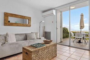 A living room with a sofa, a woven coffee table, a mirror, and a sliding glass door leading to an outdoor patio with chairs and an umbrella.