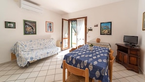 A room with a sofa, a dining table with a patterned tablecloth, a television on a wooden cabinet, and framed pictures on the walls. - La Conchiglia Apartment (Seccheto)