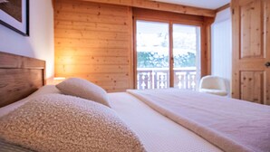 Un dormitorio con techo de madera, una cama grande con colchón blanco y dos almohadas color beige, un armario de madera y un balcón con una silla blanca.