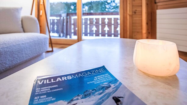 Una revista titulada 'VILLARS MAGAZINE' sobre una mesa, con una vela encendida, piso de madera y vista a un paisaje nevado a través de una ventana.