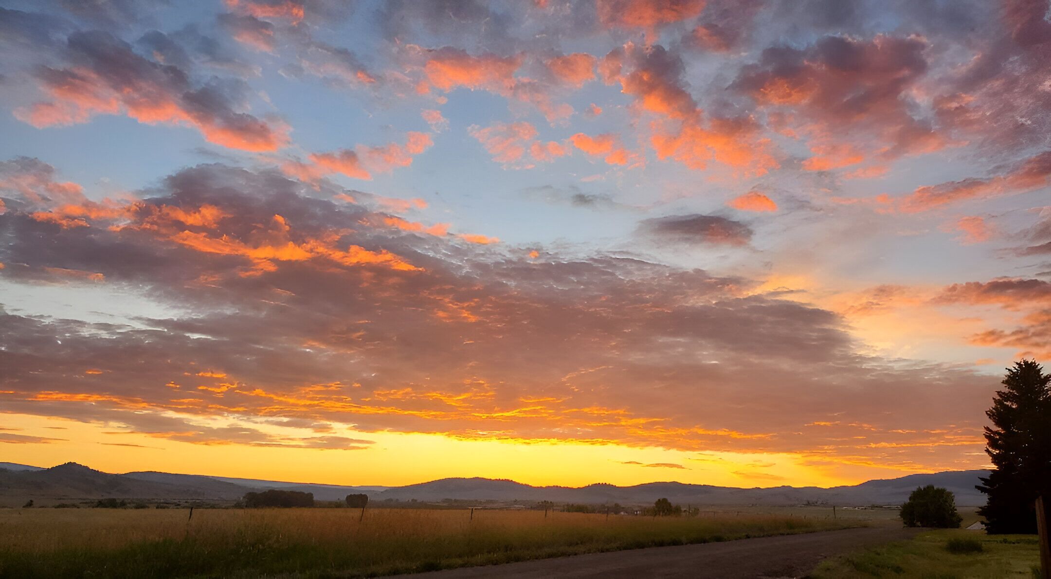 Een landelijk landschap met een onverharde weg, velden en verre bergen onder een kleurrijke zonsondergang.