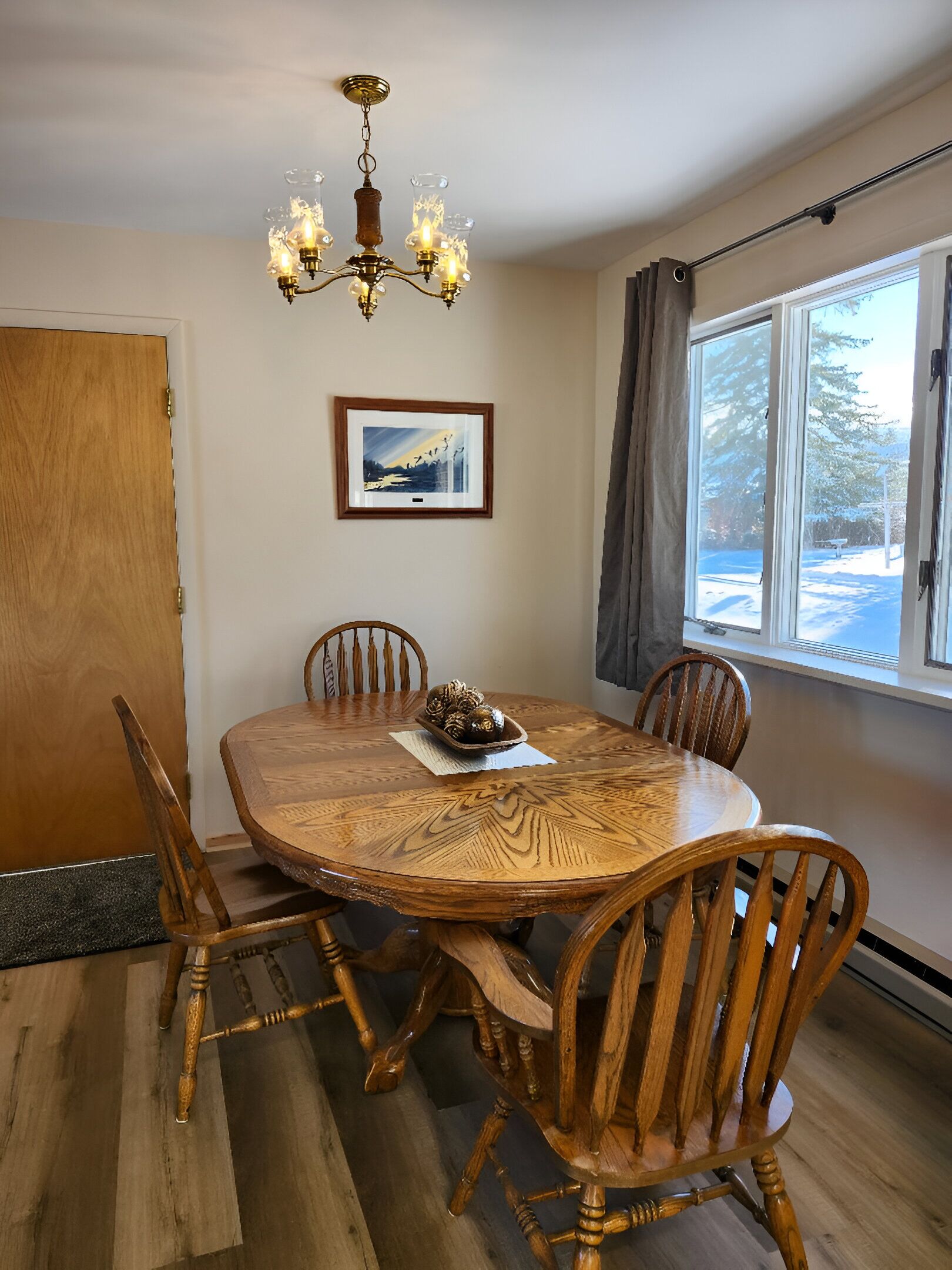 A dining room with a wooden table and chairs, a chandelier, a framed picture on the wall, and a window with a view of snow outside.