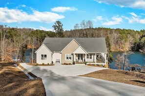Une maison à deux étages avec un porche couvert, une allée et un lac en arrière-plan.