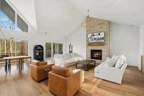 A spacious living room with a fireplace, white sofas, leather armchairs, and a dining area with a long table and chairs.