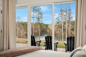 A bedroom with a large window overlooking a lake, two black chairs, and a beige bedspread.
