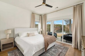 Une chambre à coucher avec un lit, des tables de chevet, un ventilateur de plafond et une vue sur un lac, visible à travers de grandes portes vitrées.