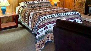 A room with wooden panel walls, a bed with patterned bedding, two bedside lamps, a framed picture on the wall, and a brick accent wall. - Northwoods (#6) Cabin in the Pines (Pinetop-Lakeside)