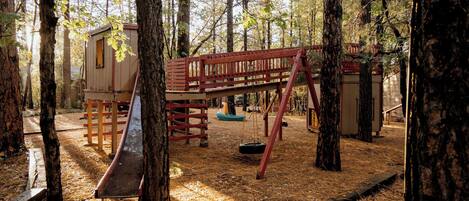 A playground with a swing set, a slide, and a small cabin-like structure surrounded by trees.