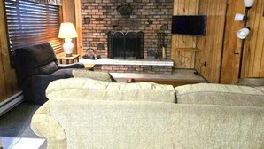 A cozy living room with a brick fireplace, wooden walls, a television mounted on the wall, and a comfortable sofa. - Northwoods #1 Cabin in the Pines (Pinetop-Lakeside)