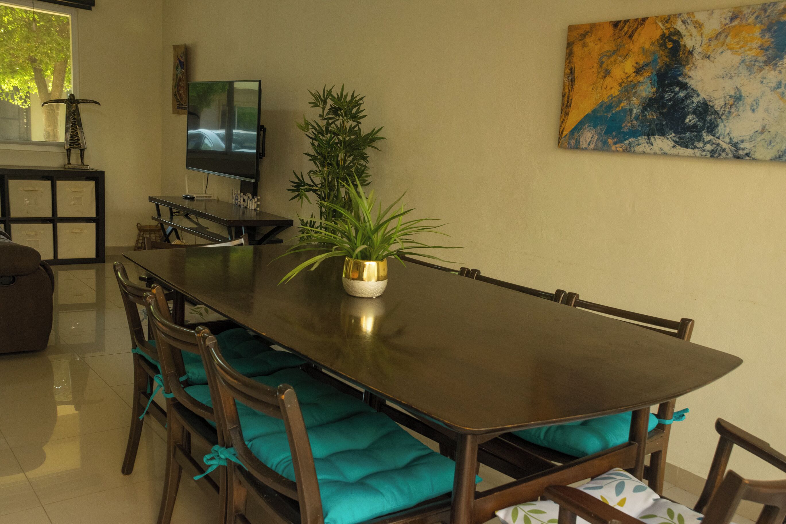 Uma sala de jantar com uma mesa comprida, cadeiras com almofadas verde-água, um vaso de planta, uma televisão e um quadro na parede.