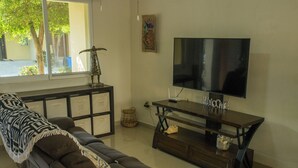 A living room with a sofa, a TV on a stand, and a window with a view of trees. - Tropical Getaway in Flamingos near Beach (Bucerías)