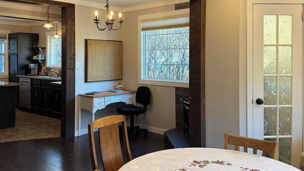 Un coin repas avec une table ronde, des chaises en bois et un lustre. On aperçoit une cuisine à l’arrière-plan, avec des meubles sombres et une fenêtre.