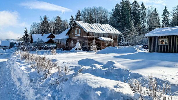 Una cabaña rústica con techo nevado y paredes de madera, rodeada por un paisaje invernal y un bosque denso.