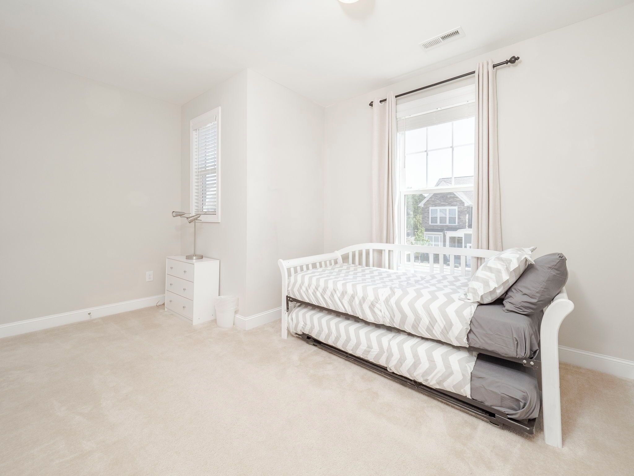 Un dormitorio con una cama blanca, una cómoda pequeña, una ventana con cortinas y vistas a una casa.
