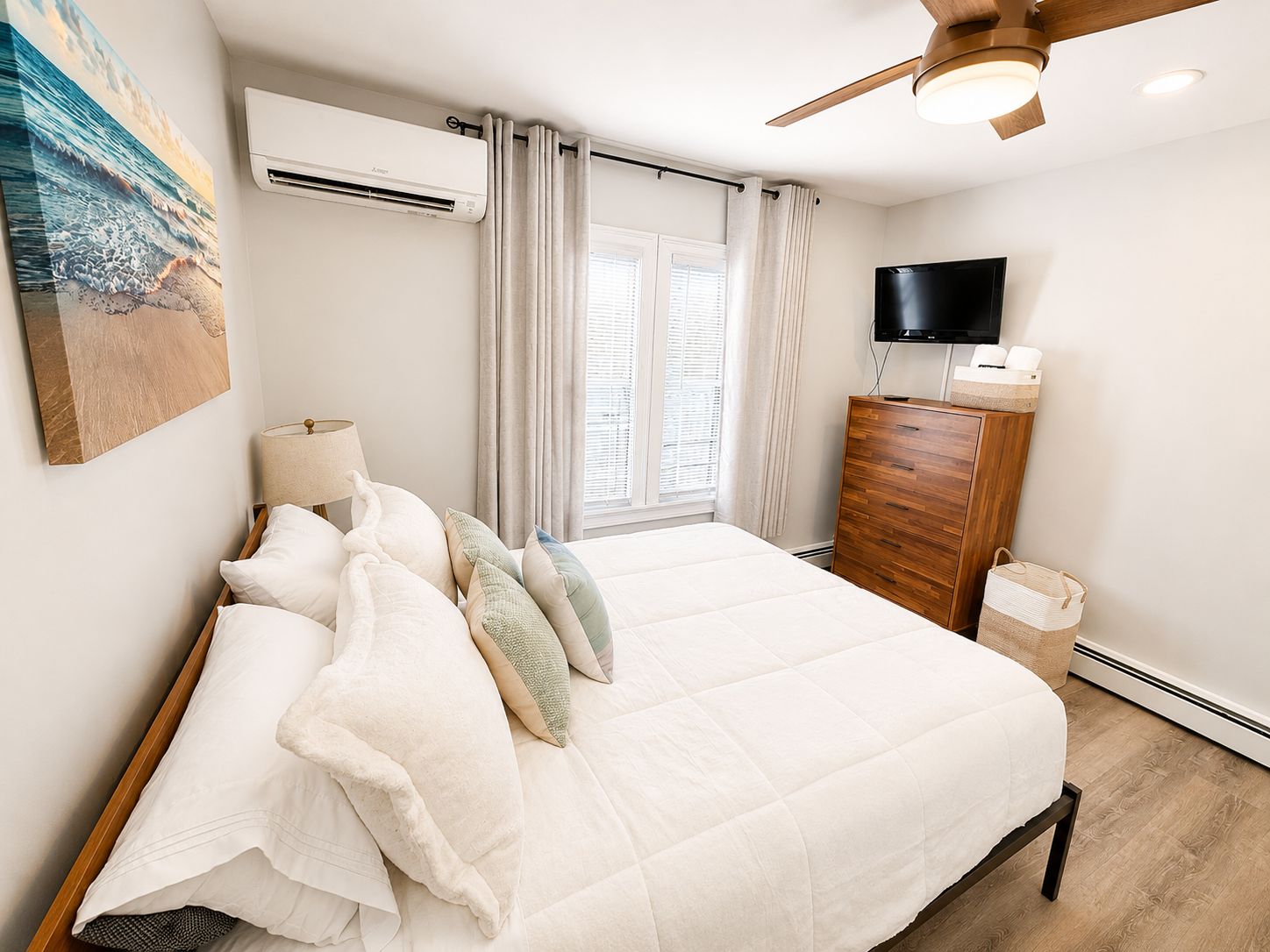 A bedroom with a bed, an air conditioning unit, a television, a wooden dresser, and a window with curtains.