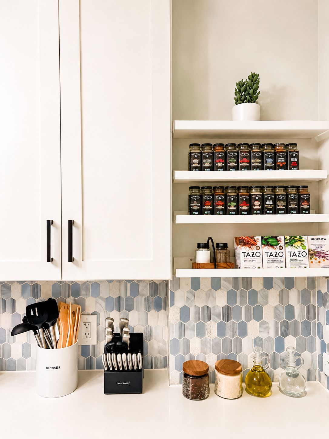 Una cocina con armarios blancos, un salpicadero de azulejos hexagonales y diversos utensilios, frascos y una planta en maceta.