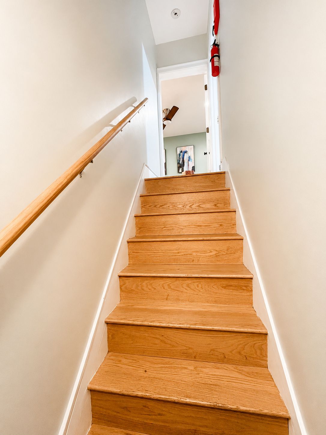 A wooden staircase with a handrail, leading to a room with a ceiling fan and a fire extinguisher mounted on the wall.