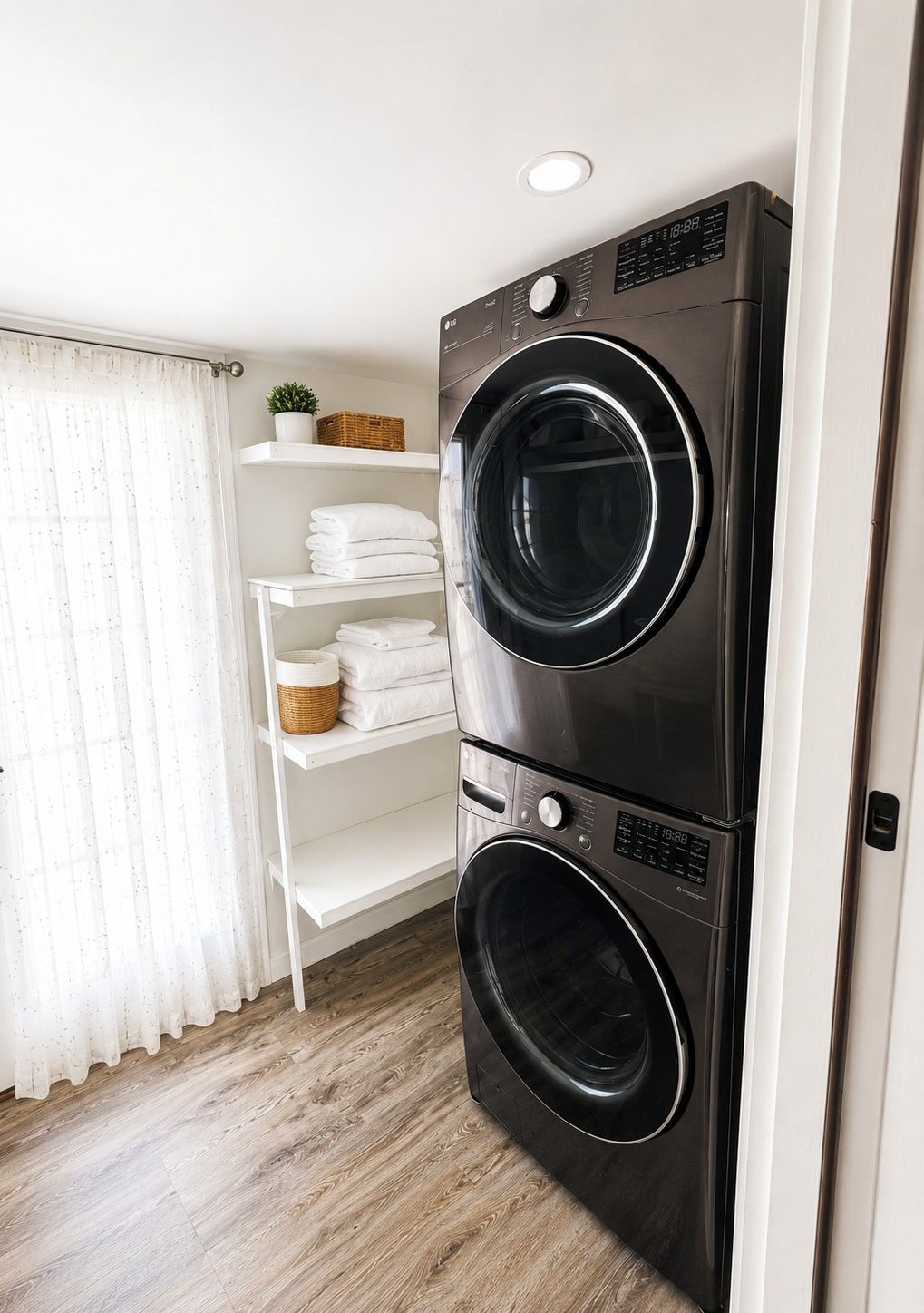 A laundry room with two stacked front-loading washing machines, a white shelving unit with folded towels, a potted plant, and a window with sheer curtains.
