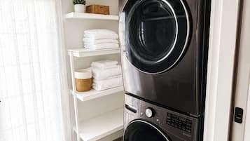 A laundry room with two stacked front-loading washing machines, a white shelving unit with folded towels, a potted plant, and a window with sheer curtains.