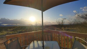 An outdoor patio with a table and chairs under a large umbrella, overlooking a desert landscape with mountains in the distance.