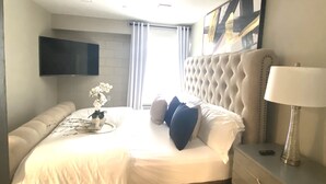 A modern bedroom with a tufted headboard, a flat-screen TV mounted on the wall, a bedside table with a lamp, and a window with curtains.