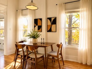 A dining area with a wooden table, four chairs, a vase with greenery, two abstract paintings, and a window with curtains.