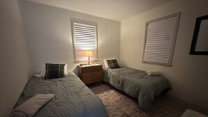 Une petite chambre faiblement éclairée, avec deux lits simples, une table de chevet et un tapis.