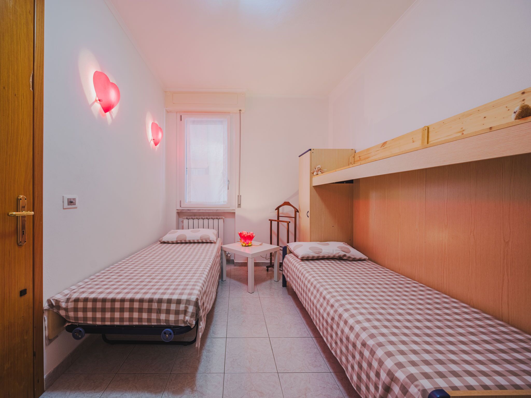 Una habitación pequeña y luminosa con dos camas individuales, una ventana, una mesa con una silla y una cama litera de madera.