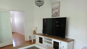 A modern living room with a flat-screen TV, white cabinets, a glass-top coffee table, and a chandelier. - Charming Queenslander in Beachmere: WiFi, A/C, Netflix, Kayo, Sleeps up to 10. (Beachmere)