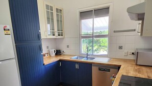 A compact kitchen with a sink, a dishwasher, and a window with a view of greenery. - Charming Queenslander in Beachmere: WiFi, A/C, Netflix, Kayo, Sleeps up to 10. (Beachmere)