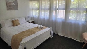 A neatly made bed with a white headboard, a bedside table with a lamp, and sheer curtains on the windows. - Charming Queenslander in Beachmere: WiFi, A/C, Netflix, Kayo, Sleeps up to 10. (Beachmere)